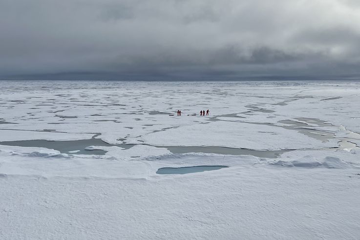 Five expedition members walk across the Arctic ice floe landscape in their red suits. 